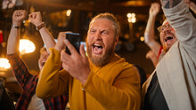 Excited Masculine Man Holding A Smartphone, Feeling Nervous About The Sports Bet He Put On A Favorite Soccer Team. Ecstatic When Football Team Scores A Goal And He Wins A High Stakes Casino Prize.