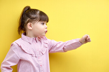 Side view portrait of child girl looking at side and speaking, points hand forward, funny and emotional kid with pony tail isolated on yellow background