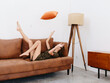 © SHOTPRIME STUDIO - Young woman lying on orange couch at home with her legs up and tossing pillow, happy relaxation at home, stylish interior, copy space