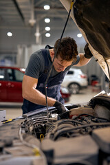 young Mid adult mechanic repairing system of car in hood in auto repair shop. view from back on caucasian guy in uniform alone in workshop car service. occupation, garage, vehicle