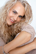 Portrait closeup of adult blonde woman with voluminous curly hairstyle hugging legs by hands on white background, copy space. Middle aged people, hair and skin care, lifting makeup, office outfit