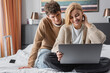 © LIGHTFIELD STUDIOS - smiling blonde woman sitting with laptop on hotel bed near young boyfriend.