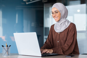 Wall Mural - Young beautiful muslim woman in hijab working inside modern office, businesswoman using laptop at work, typing on keyboard while sitting at workplace smiling and happy with achievement result.