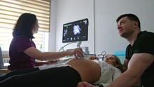 Pregnant Wife And Her Husband Are At The Doctor’s Cabinet. Female Doctor Moving The Sensor By The Big Belly Of A Woman.
