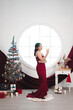© Reezky - Portrait of a pretty young girl wearing a red gown and drinking wine, smiling at the camera, standing in decorated Christmas living room indoors