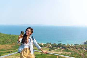 Young Asian woman traveler using mobile phone taking selfie while solo travel on tropical island mountain in summer sunny day. Attractive girl enjoy outdoor lifestyle in holiday beach vacation trip