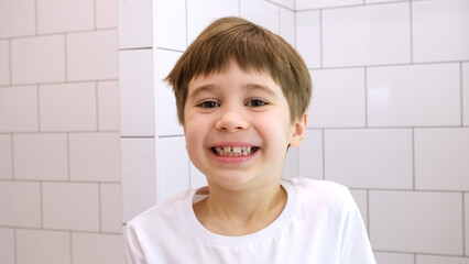 Happy child looks at the camera and smiles. Cute caucasian 6 year old boy in white t-shirt. Close-up portrait. Concept of happiness and good mood. Cheerful kid. Pretty face. Home bathroom background