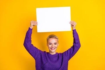 Photo of positive adorable lady wear trendy clothes arms up hold paper empty space presenting novelty isolated on yellow color background