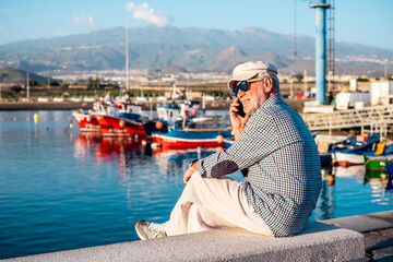 Handsome senior man in casual shirt and hat talking on phone while sitting at the port looking at boats and sea. Relaxed elderly bearded male wearing sunglasses enjoying travel or retirement