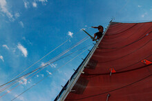 A Man Stands At The Top Of A Sailboat's Mast Near Utila Island, Honduras.