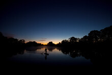 Silhouette Of One Man Stand Up Paddleboarding In Sunrise Light With Trees Reflecting In The Water.