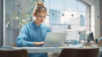 Wall Mural - Portrait of White Creative Young Woman Working on a Laptop Computer in Bright Office. Female Team Lead Smiling While Checking her Team Performance Data. Colleague Working in Background