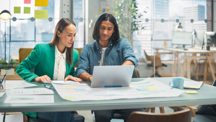 Leinwandbilder - Two Creative Colleagues Discussing and Pointing at a Laptop in Modern Casual Office. Caucasian Sales Manager Discussing Project Plan with hispanic Legal Assistant and Taking Notes