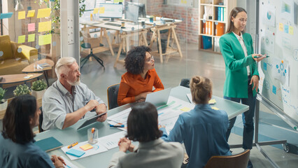 Female Company Operations Manager Holds Meeting Presentation at Office for her Team. Woman Uses a Whiteboard with Company Project Management Plan, Charts, Data. People Work in Creative Office