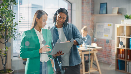 Wall Mural - Two Creative Colleagues Discussing and Pointing at a Laptop in Modern Casual Office. Female Caucasian Sales Manager Discussing Project Plan with Male Latin Legal Assistant and Taking Notes