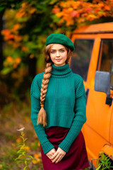 Young beautiful blonde girl with blue eyes and braided hair.Attractive model in green knitted sweater and beret standing near old retro orange bus with autumn background. Colorfull art.