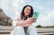 © Jose Calsina - Close up portrait of young brunette woman watching content on a cellphone app sitting outdoors. Carefree lady smiling and having fun texting messages with a smart phone. High quality photo