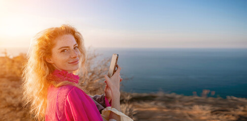 Close up portrait of curly redhead young caucasian woman with freckles looking at camera and smiling. Cute woman portrait in a pink long dress posing on a volcanic rock high above the sea at sunset