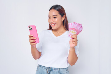 Wall Mural - Smiling young blonde woman girl Asian wearing casual white t-shirt using mobile phone and holding money rupiah banknotes isolated on white background. Fast credits concept