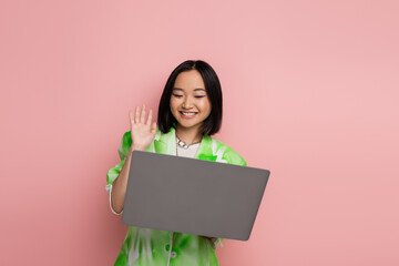 joyful and trendy asian woman waving hand during video call on laptop isolated on pink.