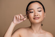 Pretty asian woman holding lush curler near face isolated on brown.