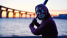 Man In The Glowing Blue Mask Threatens Waving A Bat In Hand. At Background Sunset And The Bridge At The Lake
