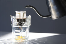Pouring Hot Water Into Glass With Drip Coffee Bag From Kettle On Light Grey Table, Closeup