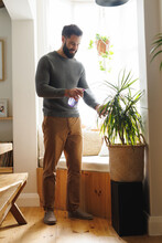 Smiling Biracial Bearded Young Man Spraying Water On Potted Plant By Window At Home