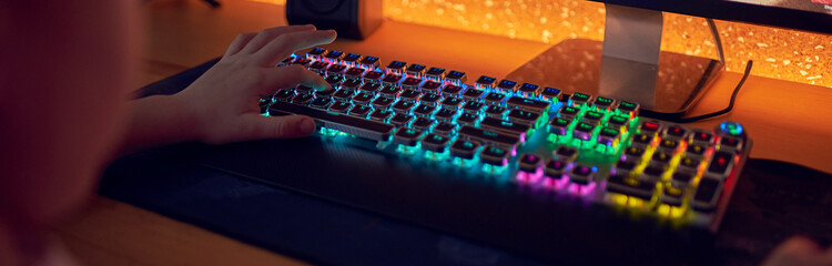 Wall Mural - Hands of teenage gamer boy playing video games on computer in dark room using backlit colorful keyboard