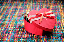 A Gift In The Shape Of A Half-open Red Heart With Dollar Notes Peeking Out Of It, On A Colourful Table.