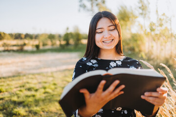 Young religious christian girl holding and reading her bible, outside in the field at sunset. Communion with God.