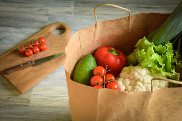 Groceries shopping concept. Closeup reusable paper bag with many vegetables fruit indoors. Cutting board with kitchen knife cherry tomatoes on background. Preparing food. Ordering product to home.