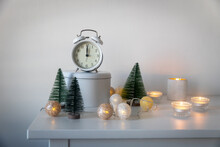 White Alarm Clock, A Twelve Pointer, A Small Artificial Green Christmas Tree And Lighted Candles In Candlesticks On A White Chest Of Drawers. Copy Space. Christmas Card.