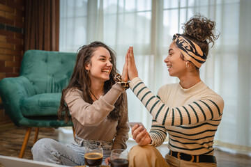 Two friends or girlfriends or sisters are high fiving and smiling because they made good purchase online while using credit card on their laptop in their apartment 