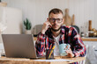 Sad Scandinavian young man in plaid shirt and glasses sits at desk with laptop holds cup of coffee with frustrated face stares away feels loneliness. Overworked caucasian guy tired. Failure, mistake.
