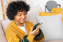 African American Woman Shopping Online Holding Smartphone Paying With Gold Credit Card. Girl Sitting At Home Buying On Internet Enter Credit Card Details. Online Shopping Ecommerce Delivery Service