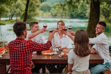 Group of happy friends toasting red wine glass while having picnic french dinner party outdoor during summer holiday vacation near the river at beautiful nature