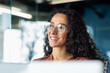 © Liubomir - Portrait of successful businesswoman wearing glasses inside office, woman looking out window smiling and dreaming happy with achievement results, Hispanic woman at workplace with laptop.