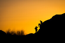Three People Silhouetted Under An Orange Sky Helping Each Other Down A Ledge.