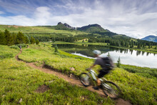 A Mountain Biker Rides By In A Blur Through Green Mountain Scenery With A Lake In The Background.