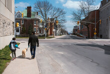 Father And Son Walking A Dog On The Sidewalk Of A Quiet City Street.