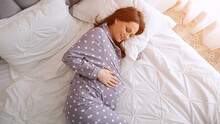 Happy Pregnant Woman Strokes Belly Lying On Cosy Double Bed. Brown-haired Mother Wearing Pyjama Waits For Baby And Takes Care Of Health, Sunlight