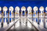 Fototapeta Sawanna - Symmetrical nightshot of the colonnade of the Sheik Zhayed mosque, with a marble catwalk surrounded by water