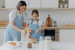 © VK Studio - Brunette mother adds oil in dough, little daughter helps to make pastry, whisks ingredients, pose together against kitchen interior, prepare bakery together. Small helper with mommy. Homemade food