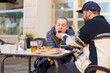 A disabled person eating on the terrace of a restaurant with a friend helping him eat