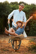 © _KUBE_ - Happy bearded father in a straw hat is pushing a cart with a cheerful little daughter. Family weekends together in the backyard. Vertical. The concept of gardening