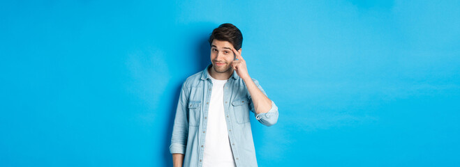 Young thoughtful man pointing at head, asking to think about it, giving a hint, standing in casual clothes over blue background