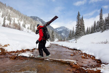 A  Man Carries His Skis Across A Snowy, Mountain Stream.