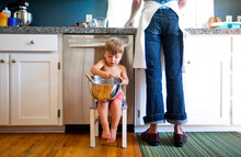 A Little Boy Sits On A Small Chair In The Kitchen And Licks The Bowl After Helping His Mother Bake A Cake.