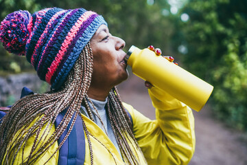 Wall Mural - Senior african woman drinking water during trekking day in mountain forest - Focus on hat - Travel and winter trekking concept